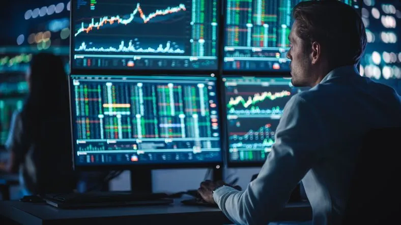 A financial analyst working at a multi-monitor workstation showing real-time stock prices.